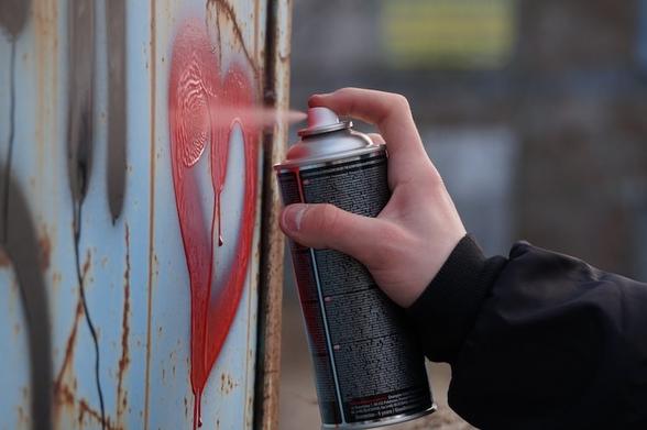A white hand holds a spray can and sprays a red heart shape on a wall