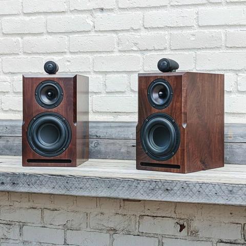 Two solid walnut speakers sit on a bench against a white brick building.
