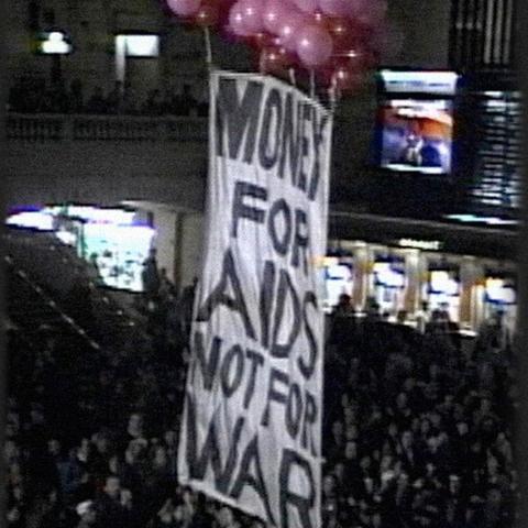 A sign that says:
Money for AIDS not for war, raised by pink and red balloons above a crowd at Grand Central terminal.
