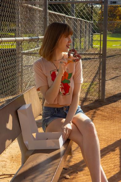Photograph of Lety seated on a dugout bench by the baseball diamond at a park. She is wearing a floral-print crop top with the text "Mexicana" on it and denim shorts. Lety's holding a heart-shaped churro dipped in chocolate and sprinkled as she stares off into the distance. She's not happy that there's no churro emoji. A donut is absolutely not the same thing. The closest Lety could get is by using Emoji Kitchen to mash-up the donut with the dumb ghost thing, but now her churro's got eyes.