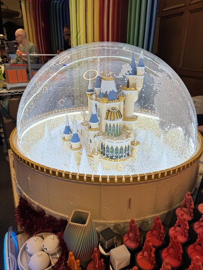 A fairytale style 3d printed castle in white, gold and blue inside a clear plastic dome, surrounded by small white polystyrene pebbles like snow