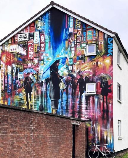 Streetartwall. This fantastic mural of a city scene in the rain and at night was sprayed/painted on the outside wall of a two-storey building. The manga-style scene of a shopping street in a large Asian city with countless illuminated advertisements, signs, pedestrians with umbrellas and a night sky looks very lively and is fantastically realized. In the foreground, a single young woman in a short black business dress stands under a blue umbrella and looks to the right. In the photo, street art photographer Tricia's bicycle is leaning against the house.