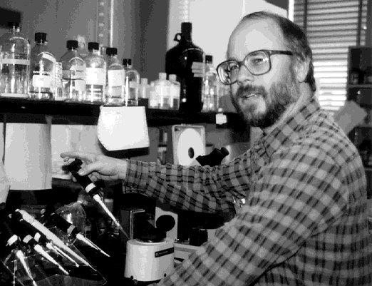 A black-and-white photo of Michael Behe. He is sitting at a biochemistry lab bench. He is holding an analytic syringe as if he is conducting an experiment, but his head is tilted to the side, awy fro mthe experiment and towards the camera. On the bench are other analytical syringes and some sort of a device. On a shelf above the bench is a row of glass bottles.