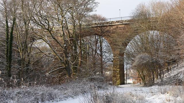 An old railway viaduct - three arches visible - stretching across a snowy landscape of wintry bare trees, snow covered land, and a grey sky behind.