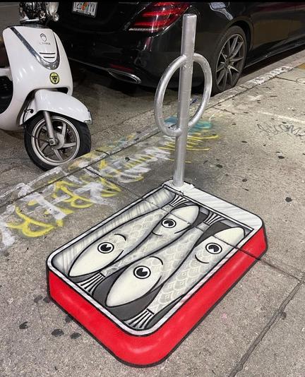 Streetart. The artist TomBob has sprayed/painted a open can of sardines on the ground next to a curved bicycle stand. The American bicycle stand is a construction of a silver metal tube in the middle and two semicircles at the top on the sides and serves here as a can opener. The red fish can is painted with a 3D effect and contains 4 cheerful fish in black and white.