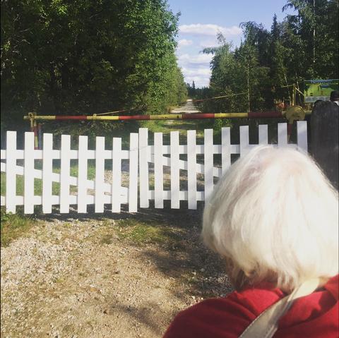 Harmaahapsinen iäkäs nainen seisoo rajavyöhykkeellä, selin kuvassa, katse kohti valkoista puuaitaa ja rajapuomia. Niiden takana jatkuu tie, jonka molemmin puolin metsää. Kesä.