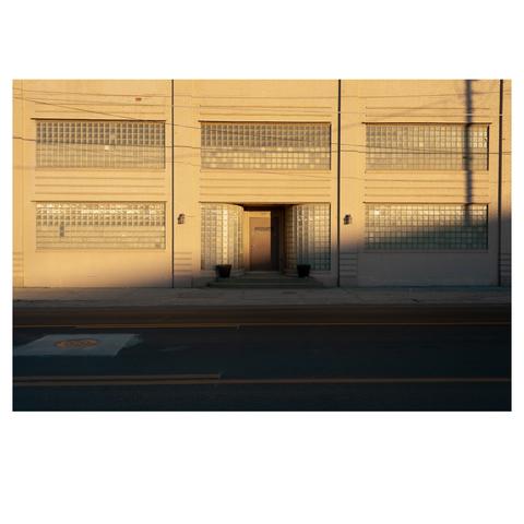 A multistory pale yellow brick building is bathed in yellow light from a setting sun. A paved street sits in front and is in shadow. In the center of the building, there is a closed door marked "private." Rectangular windows made of tiny partially opaque squares surround the door on all sides.