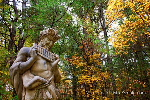 A statue of Pan with pipes in hand against an autumn forest of deciduous and fir trees.