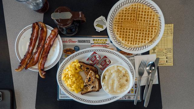 Breakfast at Waffle House that includes scrambled eggs with cheese, raisin toast, apple butter, grits, a waffle and bacon