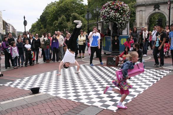 Een groep jongeren geeft een streetdance voorstelling op straat, voor de ingang van een park. Ze vertonen hun kunsten op een zwart wit stuk vinyl. Op het moment van de foto staat 1 jongen op zijn handen, een klein meisje rent voor hem langs met onder haar arm een roze speelgoed gitaar. Er staat veel publiek te kijken.
