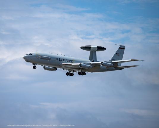 A large 4 engine military plane against a could filled sky. Atop the plane mounted on two struts is a huge saucer that houses the radar