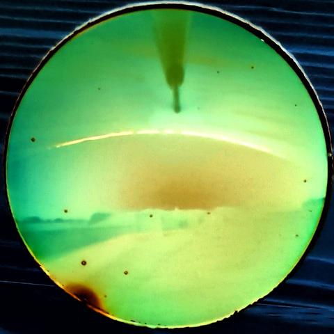 A yellow-green and green disk against a dark blue woodgrain background. In the disk is a central area of orange, and an off-white thin arc. From the top of the dis is the shadow of an anemometer arm.