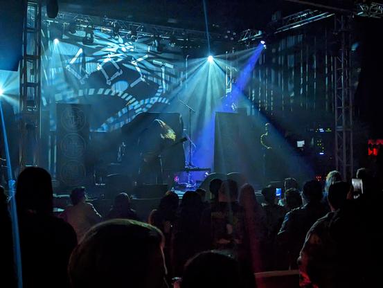 Gost on stage, dark with blue lights. Bassist to the left and keyboards to the right. The keyboardist has a skull mask