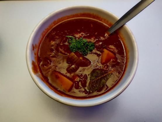 A small bowl of Goulash with a spoon in it, viewed from above.
Onion and potato chunks are just visible ind the thick soup. A bayleaf and parsley are also visible on the surface.