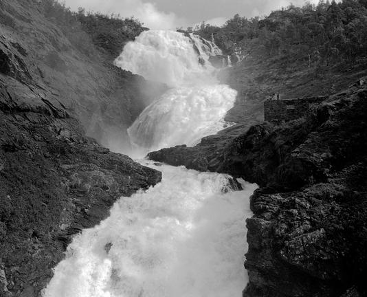 Center of frame is a waterfall, with rock on either side; right side includes the remains of a wall.