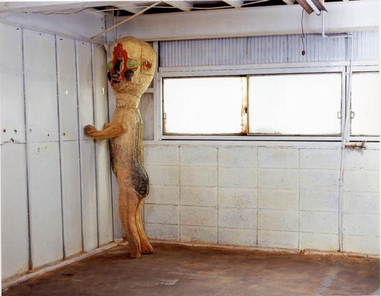 A surreal sculpture with infantile proportions and weathered, colorful paint propped against a wall in a room with dirty windows and floor.