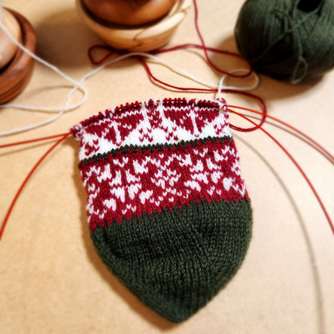 A knitted holiday stocking in progress. The toe is green, then there is a red and white pattern, followed by a white and red one. It is in the Norwegian style.