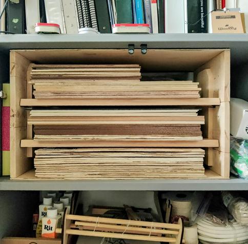 A wooden shelf holding three stacks of thin plywood sheets. In the margins, bottles of Rit dye, a small loom, and coils of cotton rope are visible.