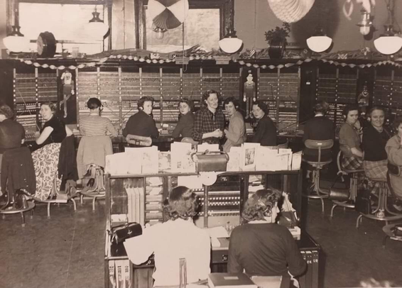 Large telephone switchboard wrapping round at least 2 walls of the room with 11 operators seated at it, 2 operators on an enquiry desk in the foreground and the exchange supervisor standing in the middle of the room.

The switchboard is adorned with paper garlands, articulated paper cutouts of santa and his elves, and concertina paper decorations hanging from the ceiling.

The enquiry desk in the foreground is topped by a large collection of christmas cards.