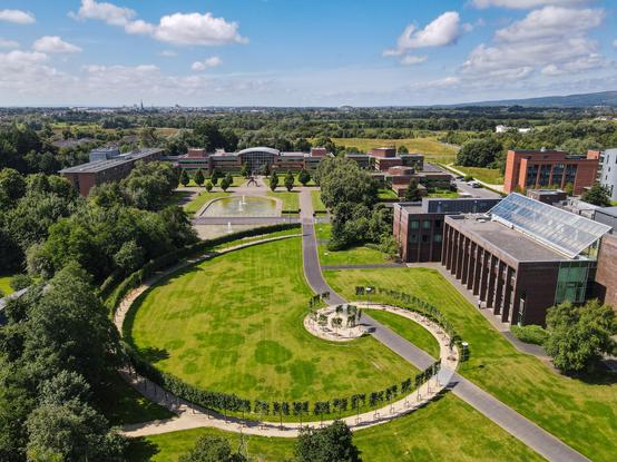 A photograph of the University of Limerick campus taken from the air. It shows a large grass lawn with a concentric circle of trees in the center. There are many brown brick buildings nestled amongst dense, mature trees. The sky overhead is bright and blue with fluffy clouds