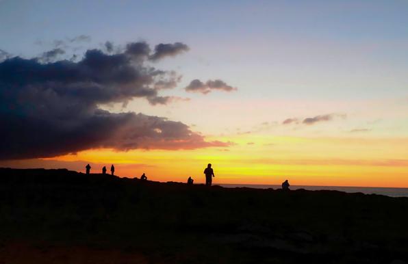 An evening sky that glows reddish and orange in the last light of day. On the horizon you can see the waterline of the ocean, in front of it, against the light, on a rocky surface, seven visible figures looking out to sea and enjoying the sunset. A large dark cloud enters the scene from the left.