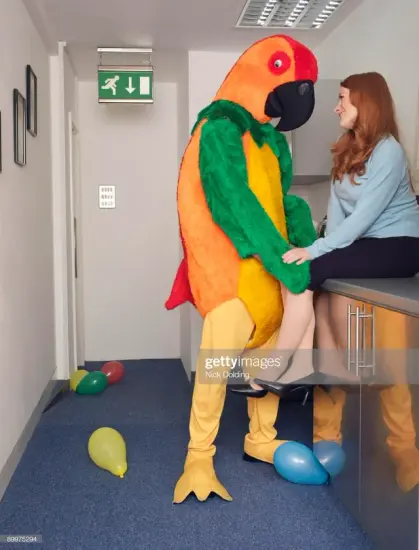 [Getty Images Stonk Pic]
A person in a colorful parrot costume perched up in a what appears to be an office hallway flirting up a white female redhead seated on a countertop, surrounded by random balloons scattered all over the floor.