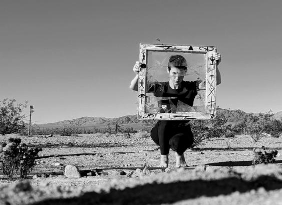 This photo is in black & white. In the foreground, rocks and marigold flowers dot the landscape. In the background a mountain juts up out of the valley, creosote and date palms dot the midground. The focus is a crouching woman with short hair holding up a broken window in front of her face. Her features are distorted by the shards of glass creating odd angles with one another. The frame is painted with sigils and the look on the woman's face is intense and determined.