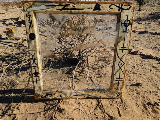 A broken window sits on the ground, a creosote bush extends branches through it. Pieces of glass are attached in places that they wouldn't naturally be, including the frame and creating odd angles with broken pieces still embedded in the frame corners. The exterior has been coated with liquid latex and is yellow-whitish, creating a strange effect that almost seems to add age to the already old reclaimed window frame. The frame is painted with various sigils all the way around. Pieces of glass have rusty red on them that appears to be blood.