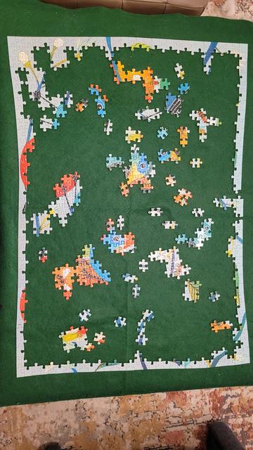 Photo of a partially finished jigsaw puzzle from above. It rests on a green felt background. The frame is almost entirely done and several chunks have been connected inside the border at various places. Shapes of birds, bees, blossoms, buds, leaves, and oranges coming