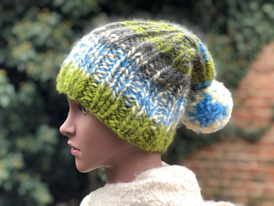 Eine Hand gestrickte Pudelmütze in den Farben, grün, hellblau, weiß und grau. Die Mütze hat eine große Bommel in weiß und hellblau.