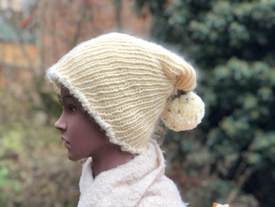 Eine Hand gestrickte Pudelmütze in Naturfarben mit einer großen Bommel in Natur,farbe und kleinen grauen Punkten