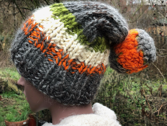 Eine Hand gestrickte Pudelmütze in den Farben, orange Schweiß und grün. Auch etwas grau ist dabei.