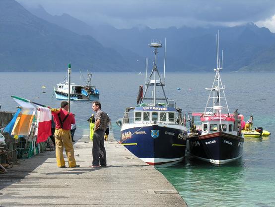 The Black Cuillin, seen under menacing clouds from the pier at Elgol on the Isle of Skye. The image shows the mountains in the distance, with the pier in the foreground with three fishing boats. There are two fishermen standing and talking on the pier. The foreground is in sunlight.