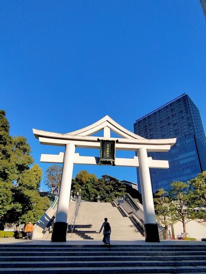 A massive white shrine gate in front of a massive set of stairs leading up the hill to the actual shrine. A woman is walking through the gate towards the stairs. Some of the city's buildings have crept into the shot as well. The sky is blue.
