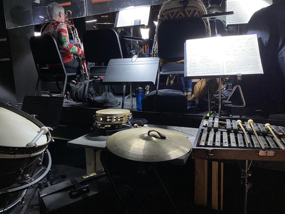 A whole mess of orchestral percussion instruments in an orchestra pit, from the player’s perspective. Bass drum, couple tambourines, some cymbals, a triangle, glockenspiel, and assorted sticks