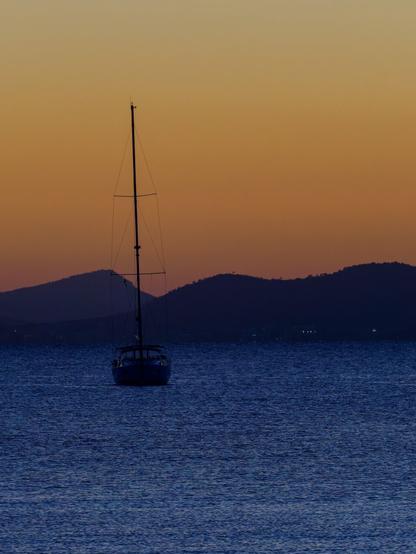 Sunset over a small sailboat anchoring before the coast with the mountains in the background. The sunset is deep orange and a nice contrast to the dark blue water.