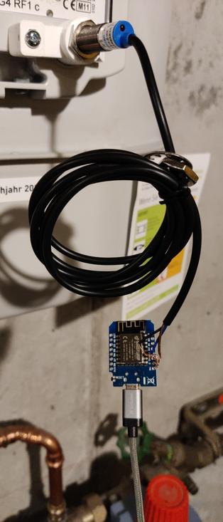 Ein Näherungssensor ist mit Hilfe eines weißen, 3D-gedruckten Teils an einem Gaszähler befestigt. An seiner im Originalzustand aufgerollten Leitung hängt, von drei gefährlich dicht nebeneinander liegenden, langen blanken Kupferdrahtenden gehalten, ein D1 mini, in dem ein ebenfalls frei baumelndes USB-Kabel steckt.