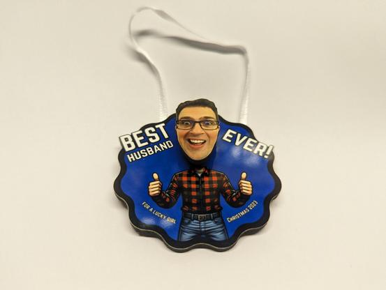 A handmade Christmas ornament. It looks like a bobblehead. There is a man's body in blue jeans and a red plaid shirt. The man's face is cut out and attached to a spring, which is glued to the background. The text says Best Husband Ever! For a lucky girl - Christmas 2023.