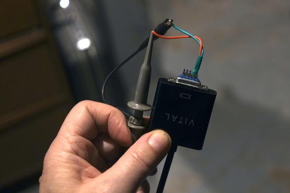 a hand showing connections between an oscilloscope probe and a DB15 connector plugged in a HDMI to VGA adapter. Only two wires, red and green, are soldered to the bare connector.