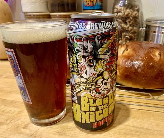 A glass of red ale and its can on a maple wood table. The can has a cartoon of a bloody unicorn.