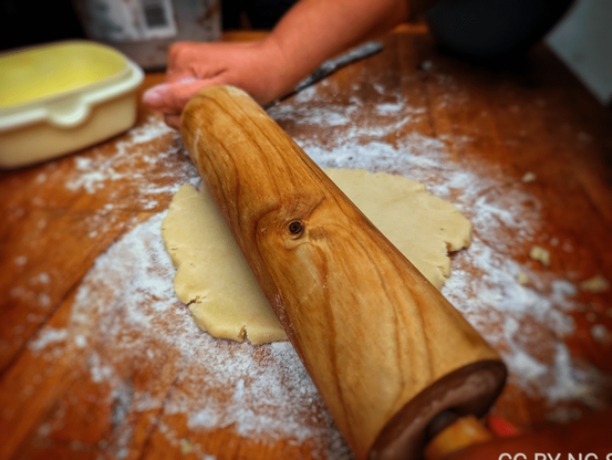 Eine mit Mehl bedeckte Arbeitsfläche, darauf ein heller Plätzchenteig, welcher mit einer Teigrolle aus Holz bearbeitet wird.

A work surface covered with flour, a light-colored cookie dough on top, which is worked with a wooden rolling pin.