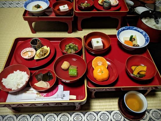 Two square red lacquer trays with red lacquered bowls with rice, pickles and assorted foods, tofu, orange, kabocha squash, mushrooms