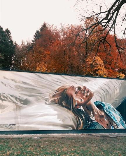 Streetartwall. The impressive mural of a blonde woman has been sprayed/painted on a long outdoor exhibition wall. In the style of an oil painting, the upper body of a young blonde woman can be seen resting her head on the edge of a bed with white sheets and looking upwards. She is wearing a light blue open blouse and her gaze seems strangely empty. On closer inspection, the shadows and highlights of her face are thin, puzzle-like lines interrupted by single broader turquoise strokes. The street wall stands in front of a row of autumnal deciduous trees. 
Info: To explain the mural, the artist says: "In the quiet expanse of her isolation, a woman lies like a living painting on the bed of her loneliness. The pixels of social distancing draw invisible lines between people and erect boundaries in a fleeting virtual world."