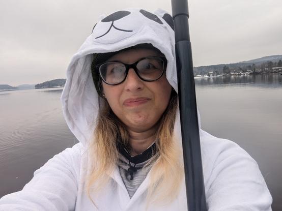 Me from the chest up wearing polar bear hooded pajamas over my clothes. The hood is up so the polar bear's face is visible. I'm holding my paddleboard paddle vertically next to my head. The lake is in the background.