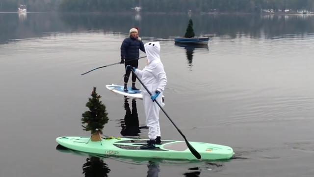 Two paddleboarders on a lake, one wearing a polar bear costume and the other in a Santa hat. One board has a small Christmas tree strapped on the front. Behind is a rowboat with a tree in it, anchored.