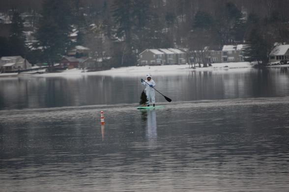 A paddleboarder wearing a polar bear costume with a small tree on the front of the board. Behind, the Lakeshore is covered with snow and there are houses.