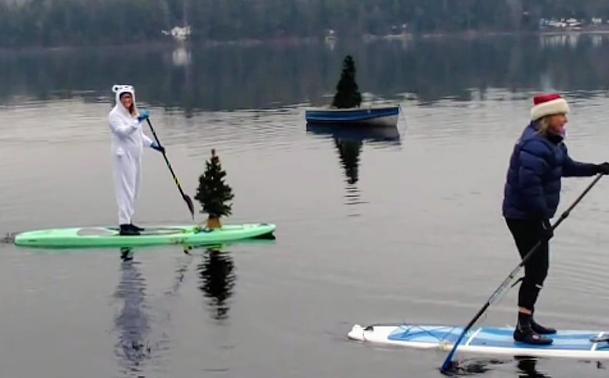 Two paddleboarders, one wearing a polar bear costume and the other in a Santa hat, paddle past an anchored rowboat with a Christmas tree standing in it.