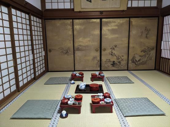 4 cushions with 4 trays sets of food on the floor in a tatami mat covered room with sliding doors on all sides. The doors in the background are a gold shade with black ink painting of trees mostly