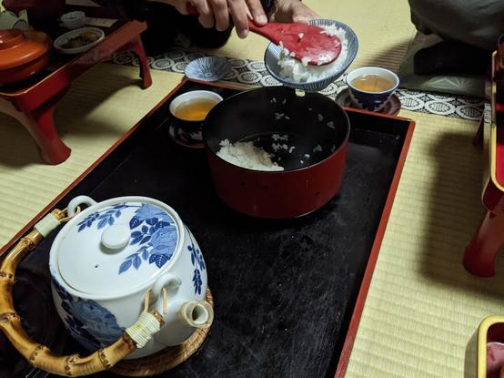 A black tray with a large white pot with blue leafy design and a wooden handle, and a red lacquered round dish that is black inside and has cooked rice in it