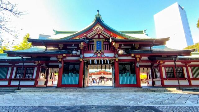 One of the side gates to the actual shrine grounds. It is itself a massive ornately worked wooden construction painted red, white, and green. It is slightly in the shade but the shrine on the inside isn't and can be glimpsed through the wide open doors at the center of this structure.